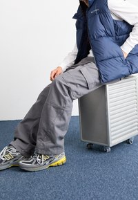 Tmavě modrá vatovaná vesta přes bílou košili s dlouhým rukávem, v kombinaci s volnými šedými cargo kalhotami a �šedými teniskami se žlutými akcenty, sedící na kovovém vozíku.