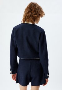 Giacca tailored blu navy in tessuto testurizzato con dettagli bianchi; design cropped, maniche lunghe e polsini con bottoni, abbinata a pantaloni corti coordinati.