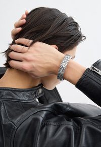 Metallarmband med rundade, präglade mönster, bärs på ett handled. I bakgrunden syns en svart läderjacka. Håret delvis täcker nacken.