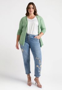 Groene textuur cardigan, witte blouse, blauwe denim jeans met borduurwerk en zilveren sandalen met bandjes.