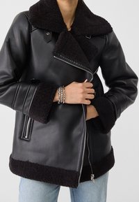 Veste en cuir noir avec un col et des poignets en fourrure noire, présentant une fermeture éclair diagonale et des poches latérales. Design texturé et chaud.