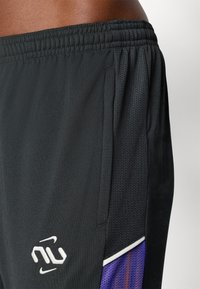 Prim-plan cu pantaloni scurți sport negri, cu panouri laterale din plasă, talie elastică și un logo brodat alb aproape de buzunar.