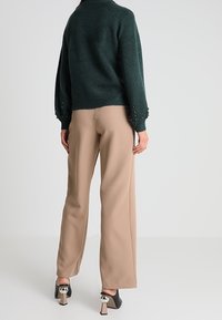 T tumma vihreä neulepaita, jossa on teksturoituja hihoja ja helmiyksityiskohtia, yhdistettynä vaaleisiin leveisiin housuihin ja mustiin korkokenkiin.