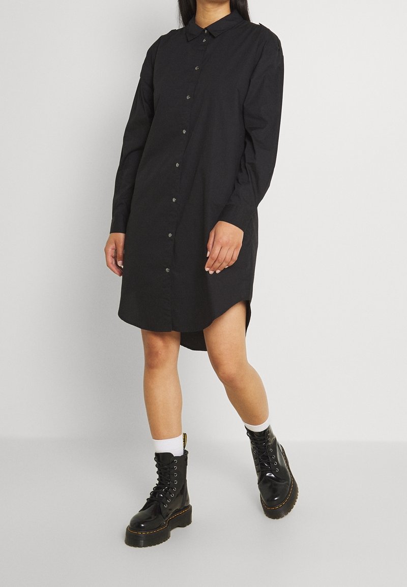 Personne portant une robe-chemise noire à manches longues boutonnée, associée à des chaussettes blanches et des bottes de combat noires à lacets.