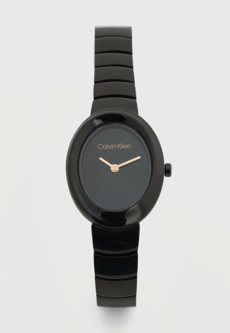 Μαύρο ρολόι χειρός Calvin Klein με οβάλ καντράν, μίνιμαλ σχεδιασμό, χρυστά δείκτες ώρας και λεπτών, και γυαλιστερό διαιρεμένο λουράκι σε ανοικτό φόντο.