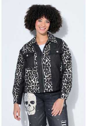 Frau mit lockigem Haar lächelt, trägt eine schwarz-weiß gemusterte Leopardenjacke und schwarze Jeans mit Totenkopf- und Farbklecks-Designs.
