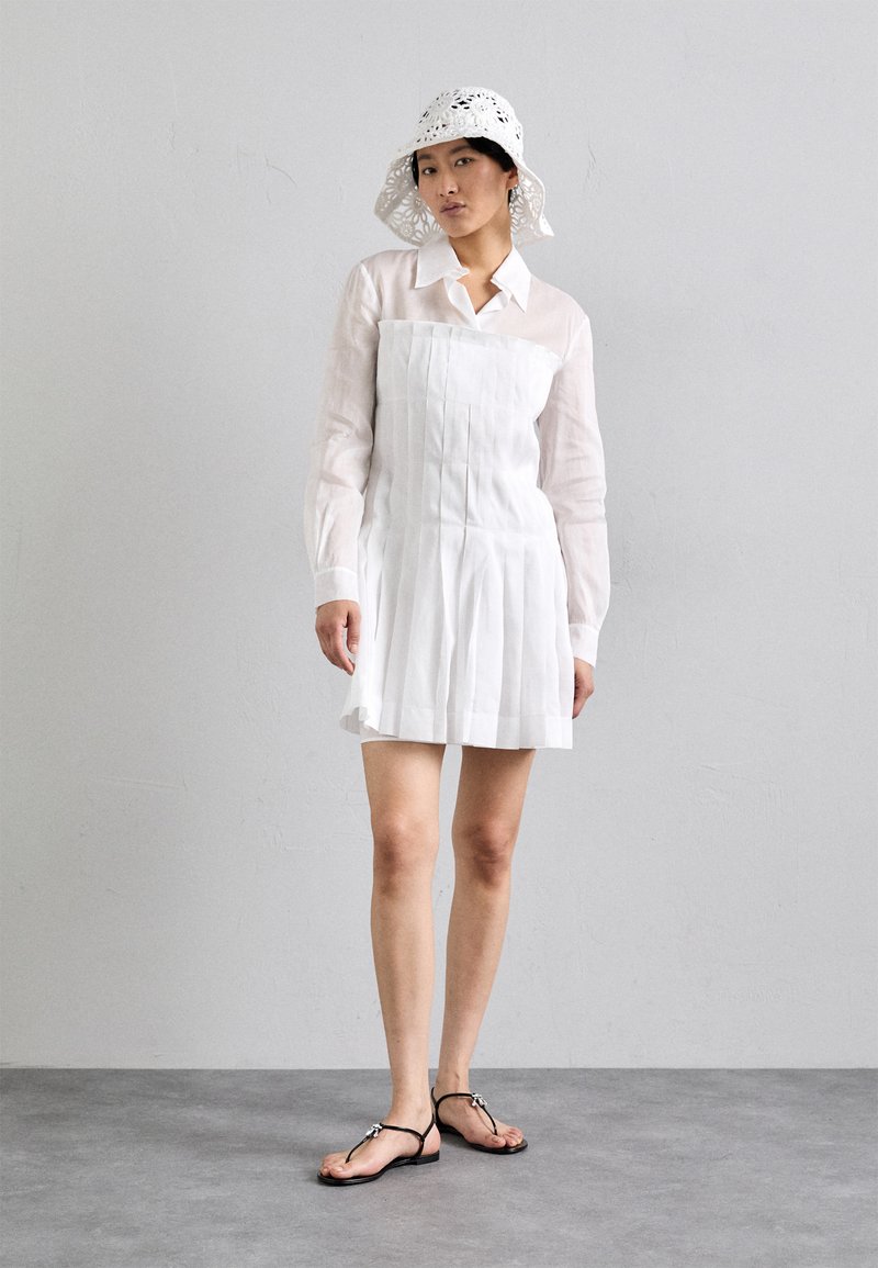 Abito bianco plissettato con design a camicia colletto, realizzato in tessuto leggero. Abbinato a sandali flat neri con infradito e un cappello a secchiello bianco in pizzo.