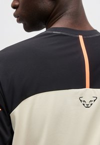 Άνδρας που φοράει μαύρο και μπεζ πουκάμισο με πορτοκαλί κάθετη ρίγα και μαύρο λογότυπο ζώου στο πάνω μέρος της πλάτης.