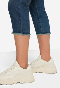 Zapatillas blancas con suela gruesa y parte superior texturizada, combinadas con pantalones capri de denim azul oscuro con dobladillo deshilachado.
