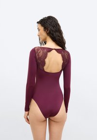 Γυναίκα με σγουρά σκουρόχρωμα μαλλιά, φορώντας ένα μακρυμάνικο μπορντό κορμάκι με δαντέλα και ανοιχτή πλάτη, στέκεται μπροστά σε ένα απλό φωτεινό φόντο.