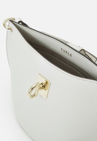 Sac à main en cuir blanc avec un accent en métal doré en forme de carré et une bague décorative. L'intérieur présente une doublure neutre avec un logo visible.