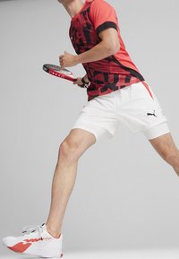 Czerwona i czarna koszulka sportowa w wzory, białe szorty i białe tenisówki z czerwonymi akcentami. Trzyma rakietę tenisową.