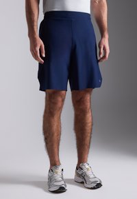 Marineblå shorts med flad talje, sidelommer og rundet kant. Båret med grå og gule træningssko, hvilket fremhæver et sporty look.