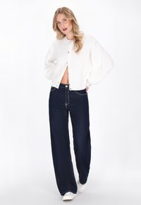 Hvid strikket cropped cardigan med guldknapper, parret med mørkeblå vide bukser og hvide sneakers, sat mod en ensfarvet baggrund.