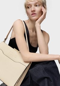 Borsa beige strutturata con logo "TOUS" in rilievo, caratterizzata da dettagli neri e una tracolla lunga. Modella che indossa un top nero e una gonna di jeans.