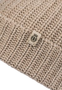 Beige knitted fabric with a small rectangular beige tag featuring a black emblem with a crown and shield design.