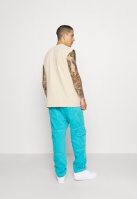 Beige ermeløs skjorte med en avslappet passform, kombinert med lyse turkise cargo-bukser med lommer og en avsmalnet bendesign. Hvite joggesko.