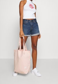 Kobieta w białym, bezrękawnikowym topie crop i dżinsowych szortach, trzymająca dużą, jasnoróżową torbę Nike, w białych trampkach, stojąca na szarym podłożu.