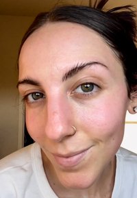 Gros plan d'un visage avec une peau claire, des yeux brun clair et des sourcils arqués. Une petite boucle d'oreille en or et un anneau nasal visibles. Texture naturelle.