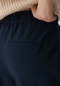 Marinblå byxor med en slät yta och elastisk midja. Har en diskret, horisontell bakficka. Diskret sömdetalj längs kanterna.