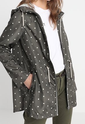 Femme portant une veste de pluie à capuche vert foncé à pois, avec fermeture éclair à l’avant et cordon de serrage, chemise blanche et pantalon vert.
