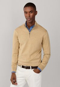 Homme portant un pull beige à fermeture éclair quart, par-dessus une chemise bleue, pantalon blanc avec ceinture noire, debout avec une main dans la poche, sur fond gris.