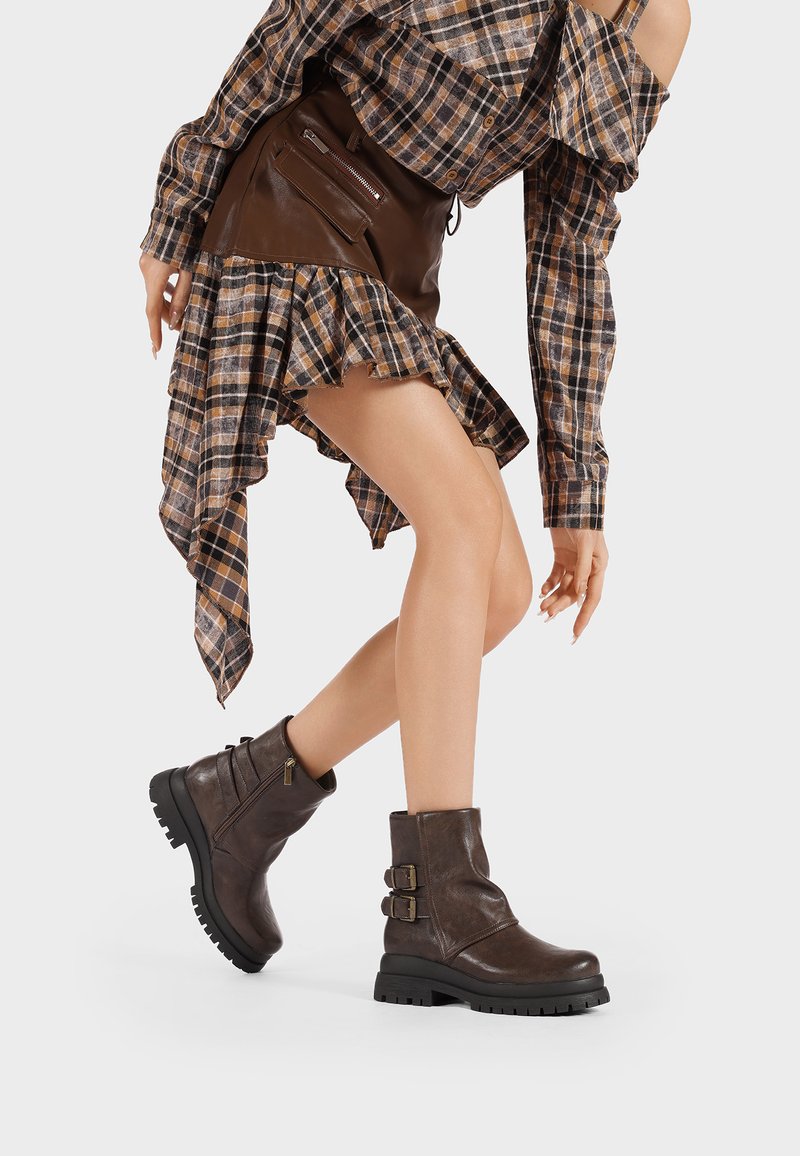 Stivaletti in pelle marrone con fibbie, finitura texturizzata, suola robusta e zip laterale, abbinati a una camicia a quadri e una gonna a strati.