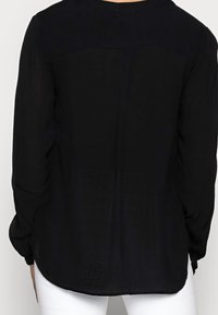 Zwarte blouse met lange mouwen, een doorzichtige achterkant en een effen voorkant, met een ronde halslijn en lichte rimpeling bij de manchetten, gemaakt van een lichte stof.