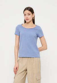 Γυναίκα με ανοιχτό μπλε κοντομάνικο μπλουζάκι και μπεζ παντελόνι cargo, στέκεται με το ένα χέρι πίσω από την πλάτη της μπροστά σε ένα απλό φόντο.