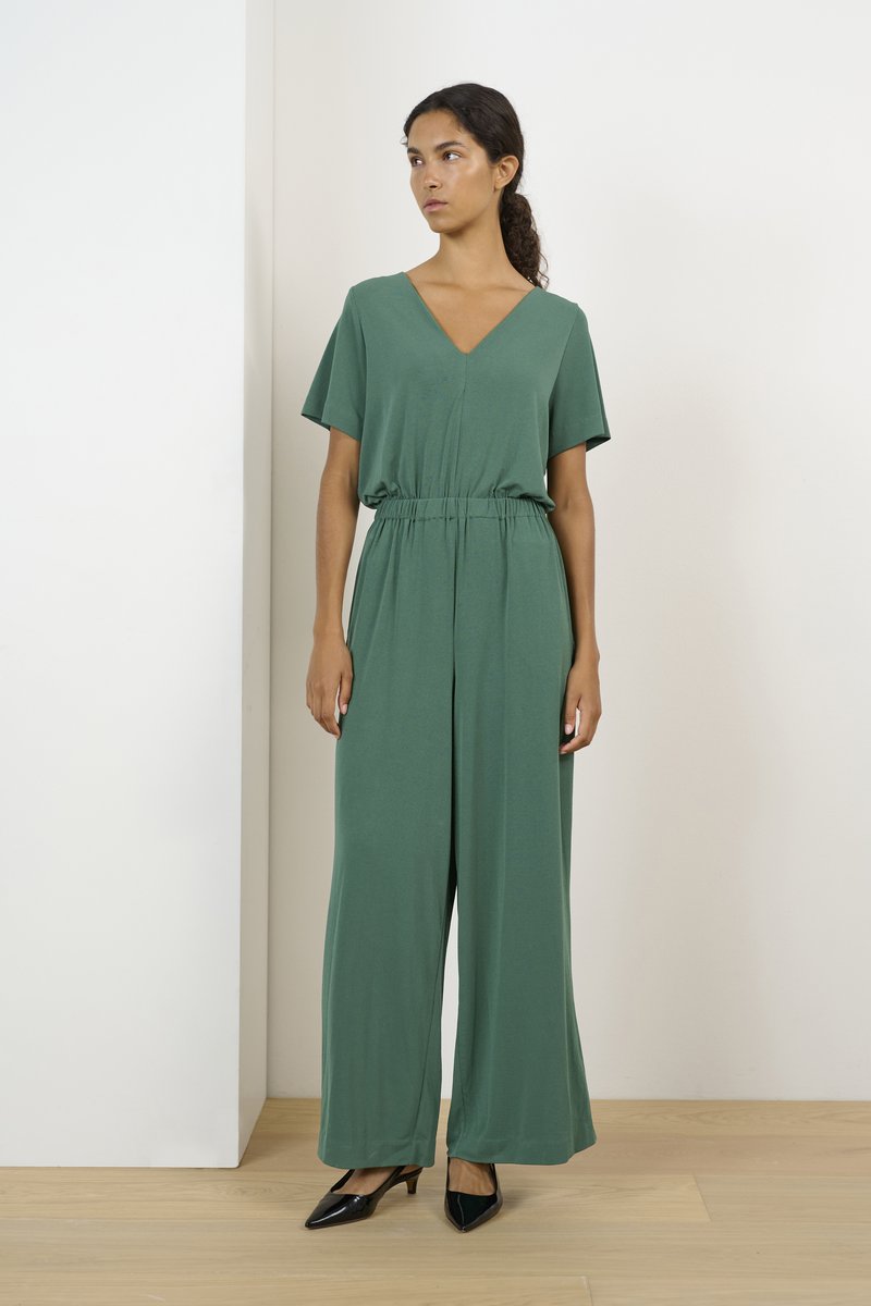 Mono verde hecho de una tela suave, con escote en V, mangas cortas, cintura elástica y diseño de pierna ancha. Llevado con tacones negros.