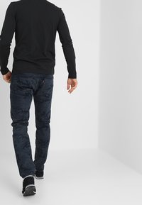 Mörkblå kamouflagejeans med avslappnad passform, parat med en svart långärmad skjorta och svarta skor, sedd bakifrån.