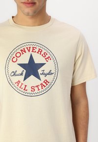Camiseta de algodón beige con un gráfico circular que presenta una estrella azul, "Converse" y "All Star" en rojo, y "Chuck Taylor" en azul escrito.