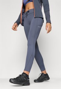 Pelēkas sporta leggingas ar pieguļošu dizainu, ar kontrastējošām šuvēm un mazu logo. Saskaņotas ar melnām taku skriešanas kurpēm.