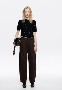 Suéter negro de manga corta con intrincados bordados florales en verde y blanco, combinado con pantalones marrones holgados y botas de tacón negras.