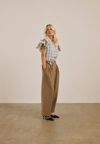 Femme portant un chemisier à carreaux à volants, rentré dans un pantalon marron taille haute à jambes larges, ceinturé d'une ceinture florale et des escarpins noirs à bout pointu.