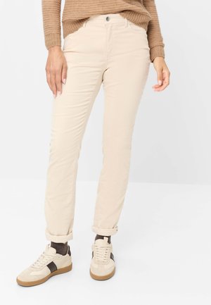 Femme portant un pantalon beige retroussé, des chaussettes marron, des baskets beige et marron, et un pull en tricot marron avec les manches baissées.