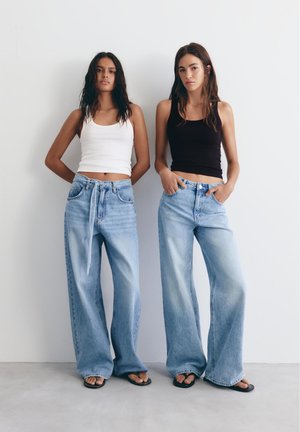 Dvě ženy stojí vedle sebe, oblečené v nátělnících bez rukávů — jedna bílá, druhá černá — a širokých světlemodrých džínách s sandály.