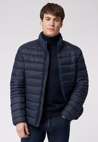 Navyblaues Puffer-Jacke mit vertikalem Steppmuster, hohem Kragen und Frontreißverschluss. Trägt man über einem dunklen Strickpullover.
