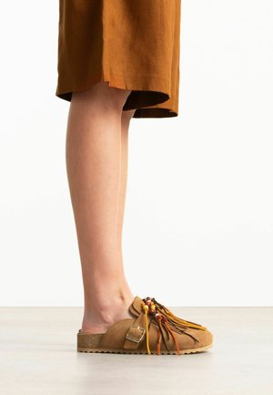 Beine, die einen braunen Rock und braune Slip-On-Schuhe mit Schnalle, Fransen und bunten Perlenverzierungen tragen, stehen auf einem hellen Boden.