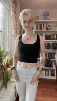 Person mit kurzem blonden Haar und Tattoos trägt ein schwarzes Crop-Top und weiße Hose, steht in einem Raum mit einem Bücherregal und Pflanzen am Fenster.