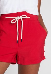 Shorts rossi a vita alta con una cintura bianca a stringa, indossati con un top senza maniche bianco a costine, con una mano appoggiata nella tasca laterale.