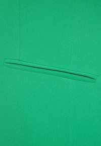 Zielony materiał w zbliżeniu, pokazujący poziomą kieszeń z porządnie wykończonymi szwami i gładką teksturą.