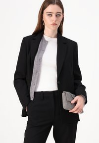 Čierny blazer s šedým kardigánom pod ním, bielym topom a čiernymi nohavicami. Drží malú károvanú kabelku s reťazovou detailom.