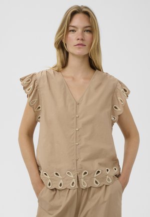 Femme aux longs cheveux blonds portant un chemisier beige boutonné avec des bords festonnés brodés et un pantalon ample assorti, les mains dans les poches.