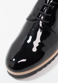 Chaussure en cuir verni noir avec une finition brillante, bout rond et design à lacets noirs. Dispose d'une semelle beige avec des accents de coutures blanches.