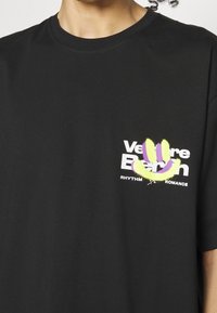 Zwart katoenen T-shirt met een kleurrafische opdruk met groene en paarse accenten, en tekst: "Venture Beach Rhythm Romance" aan de voorkant.