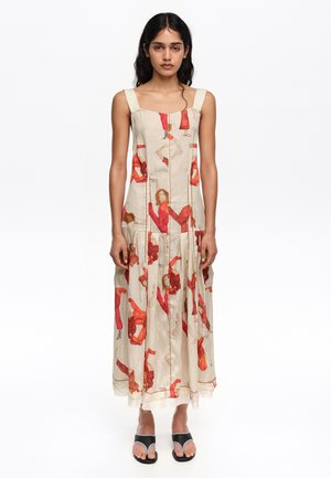 Mujer con un vestido maxi sin mangas color crema con estampados abstractos en rojo y naranja, y sandalias negras abiertas en los dedos, posando sobre fondo blanco.