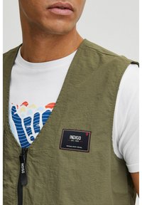 Vesta fără mâneci de culoare verde măsliniu, cu fermoar și patch brodat cu logo; purtată peste o cămașă albă ce are un design grafic colorat.