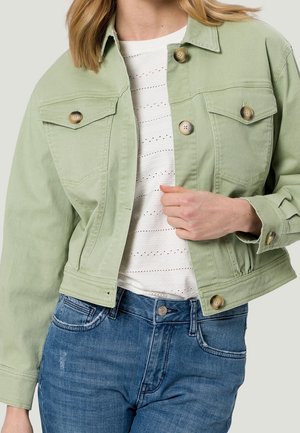 Femme portant une veste boutonnée vert clair sur un top blanc ajouré et un jean bleu, la main tenant devant la veste.