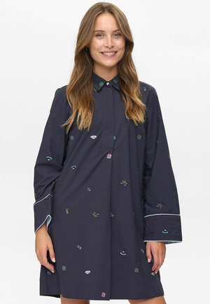 Vrouw in een losse marineblauwe jurk met lange mouwen en kleine meerkleurige geborduurde patronen, glimlachend, met los golvend haar.